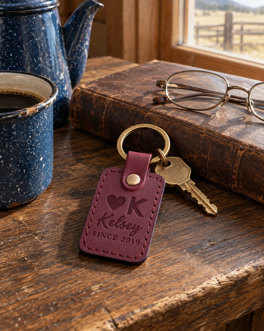 Kelsey SINCE 2019 keychain in sangria leather on a dark oak ranch kitchen table beside a speckled enamel coffee pot