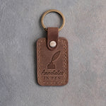 Sicilian brown leather keychain debossed with a quill in an inkwell above Annotates and IN PEN, on gray concrete