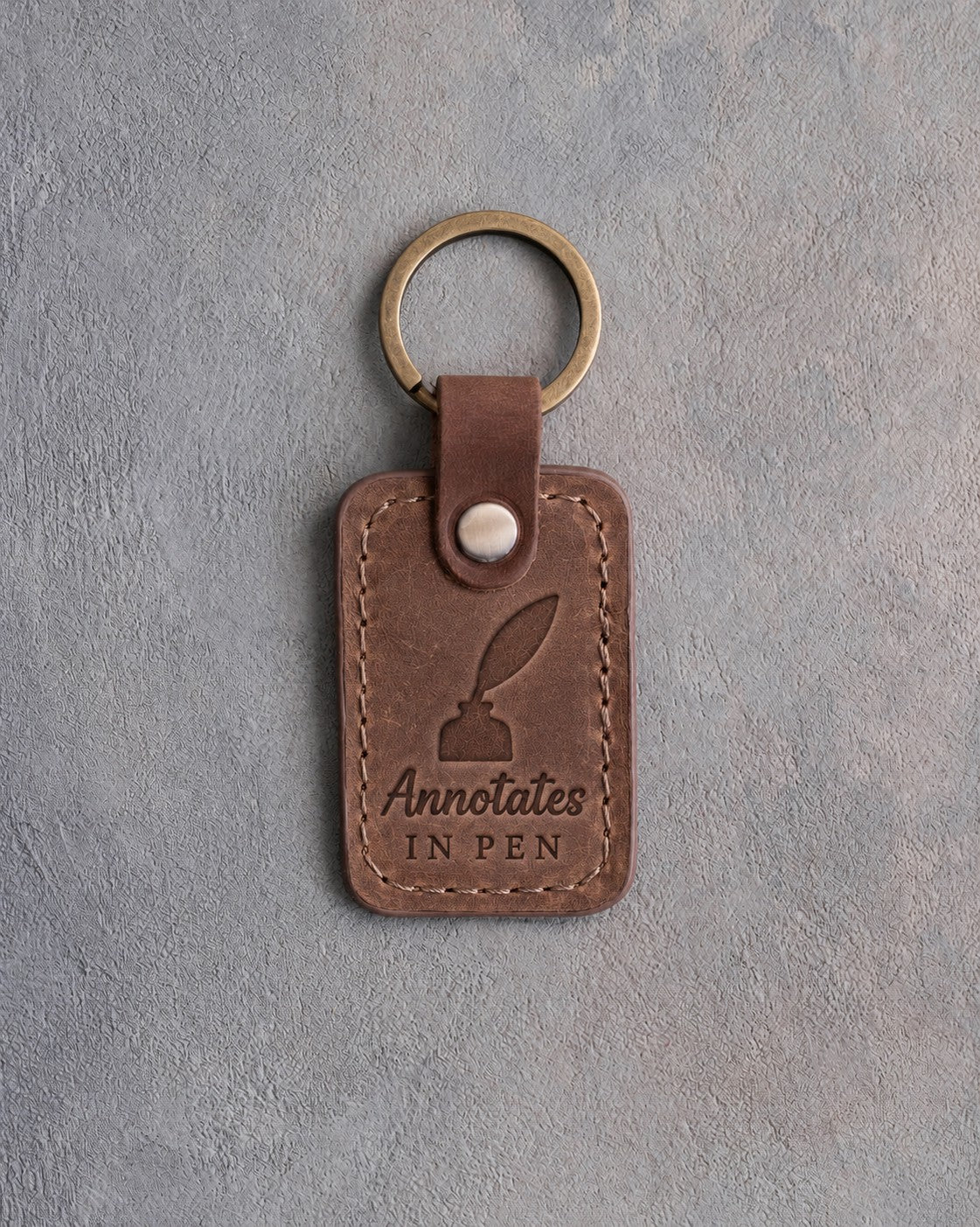 Sicilian brown leather keychain debossed with a quill in an inkwell above Annotates and IN PEN, on gray concrete
