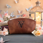 Caramel leather makeup pouch debossed with a floral letter V, set on a slate table with pale roses
