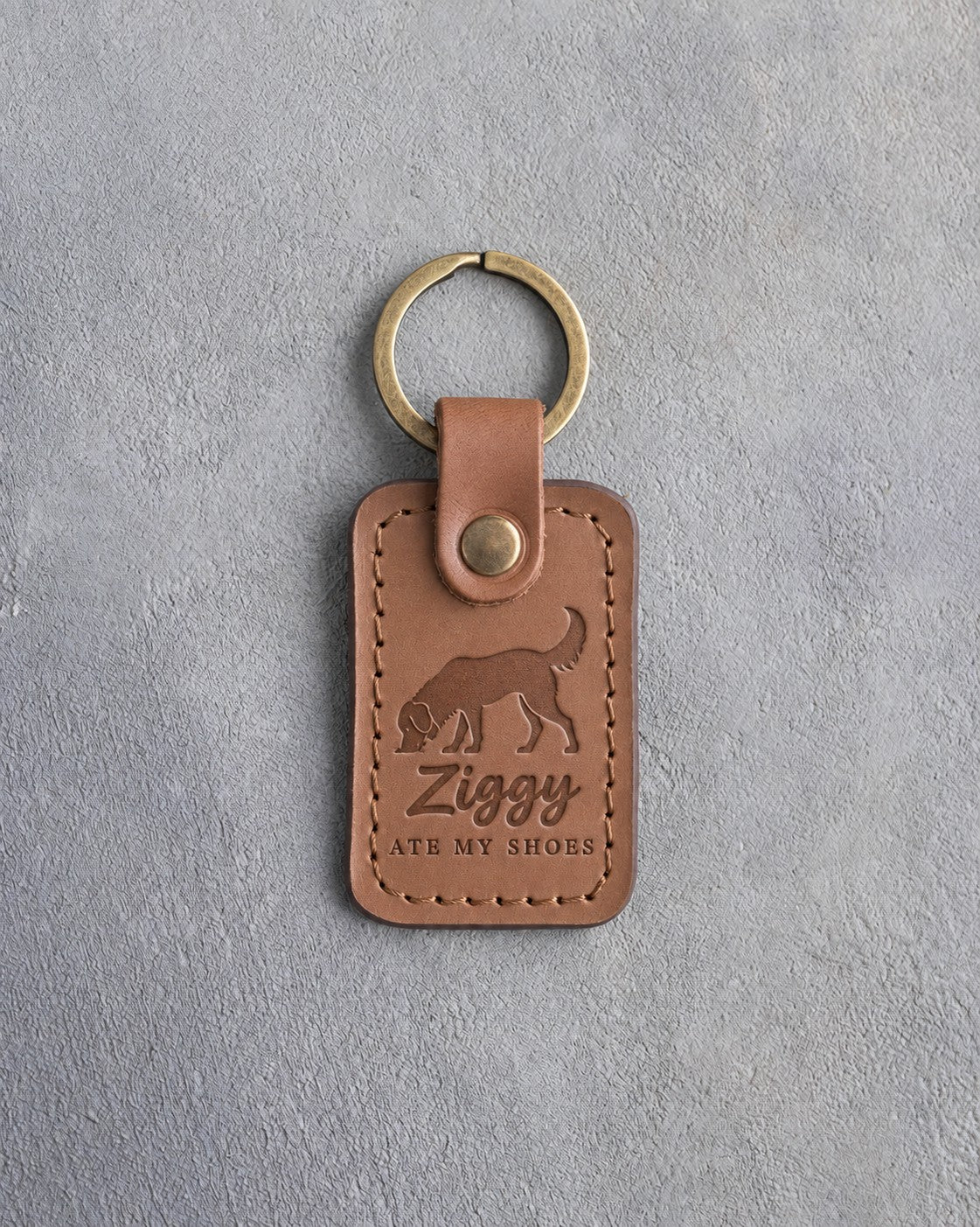 Caramel leather keychain debossed with a dog sniffing the ground above Ziggy and ATE MY SHOES, on gray concrete