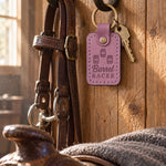 Lilac Barrel RACER keychain hanging from a forged bridle hook on a tack room wall beside a worn headstall