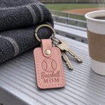 Blush Baseball Mom keychain clipped to a bat bag zipper beside a folding chair at a ballfield