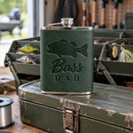 Forest green Bass DAD hip flask in full-grain leather on the lid of an olive-green steel tackle box in a garage, lures in the trays behind