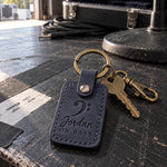 Jordan ON BASS keychain in navy leather hanging from a bass gig bag zipper beside an amplifier