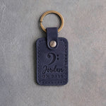 Blueberry navy leather keychain debossed with a bass clef above the name Jordan and ON BASS, on gray plaster