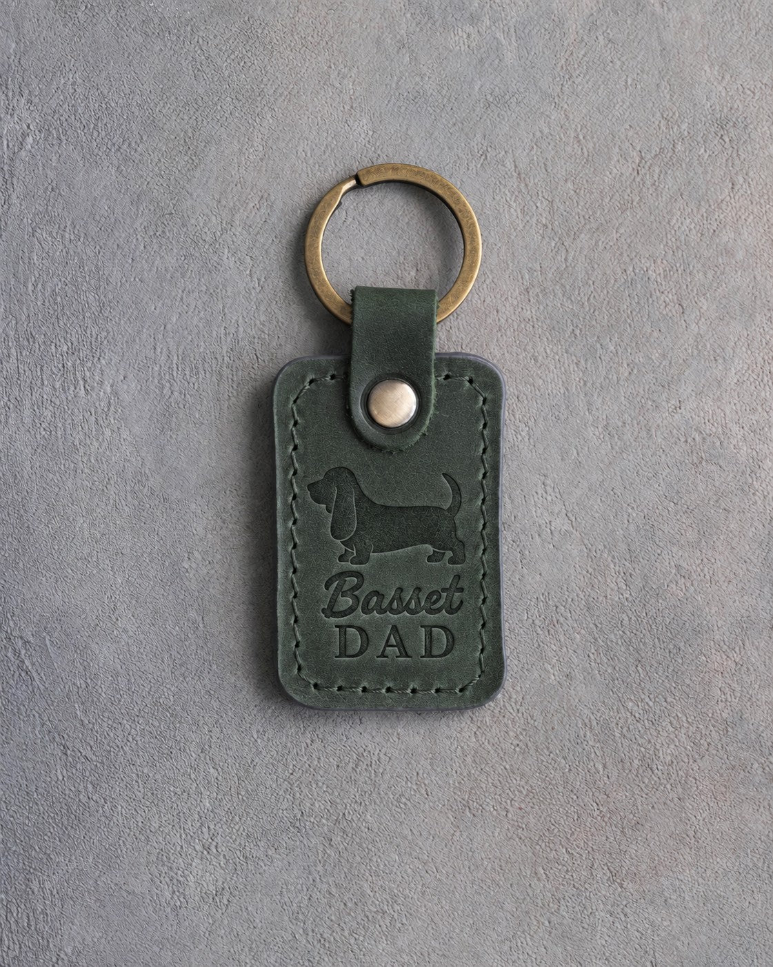 Forest green leather keychain debossed with a Basset Hound silhouette and the words Basset DAD, on gray concrete