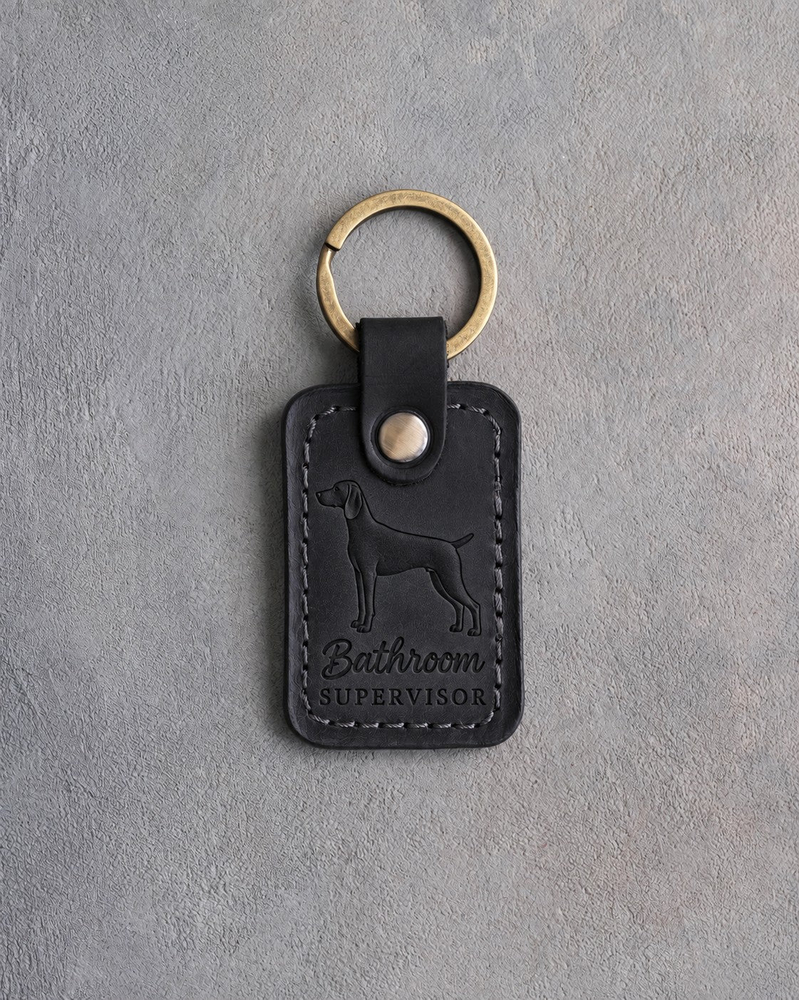 Black leather keychain debossed with a weimaraner and the words Bathroom SUPERVISOR, on gray concrete