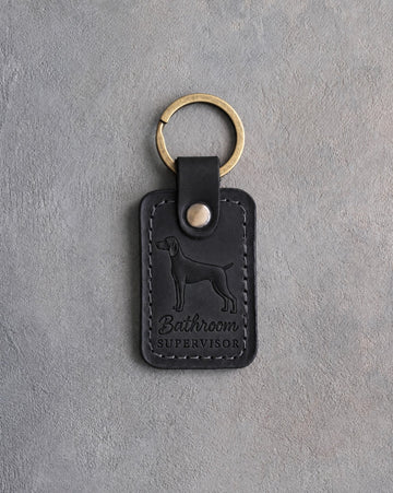 Black leather keychain debossed with a weimaraner and the words Bathroom SUPERVISOR, on gray concrete
