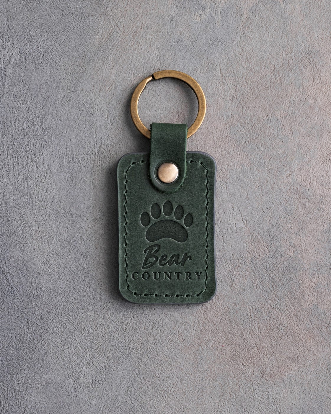 Forest green leather keychain debossed with a black bear paw track above the words Bear COUNTRY, on gray plaster