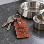 Kona BENGAL keychain in rust tan leather on a wooden shelf beside a climbing rope toy