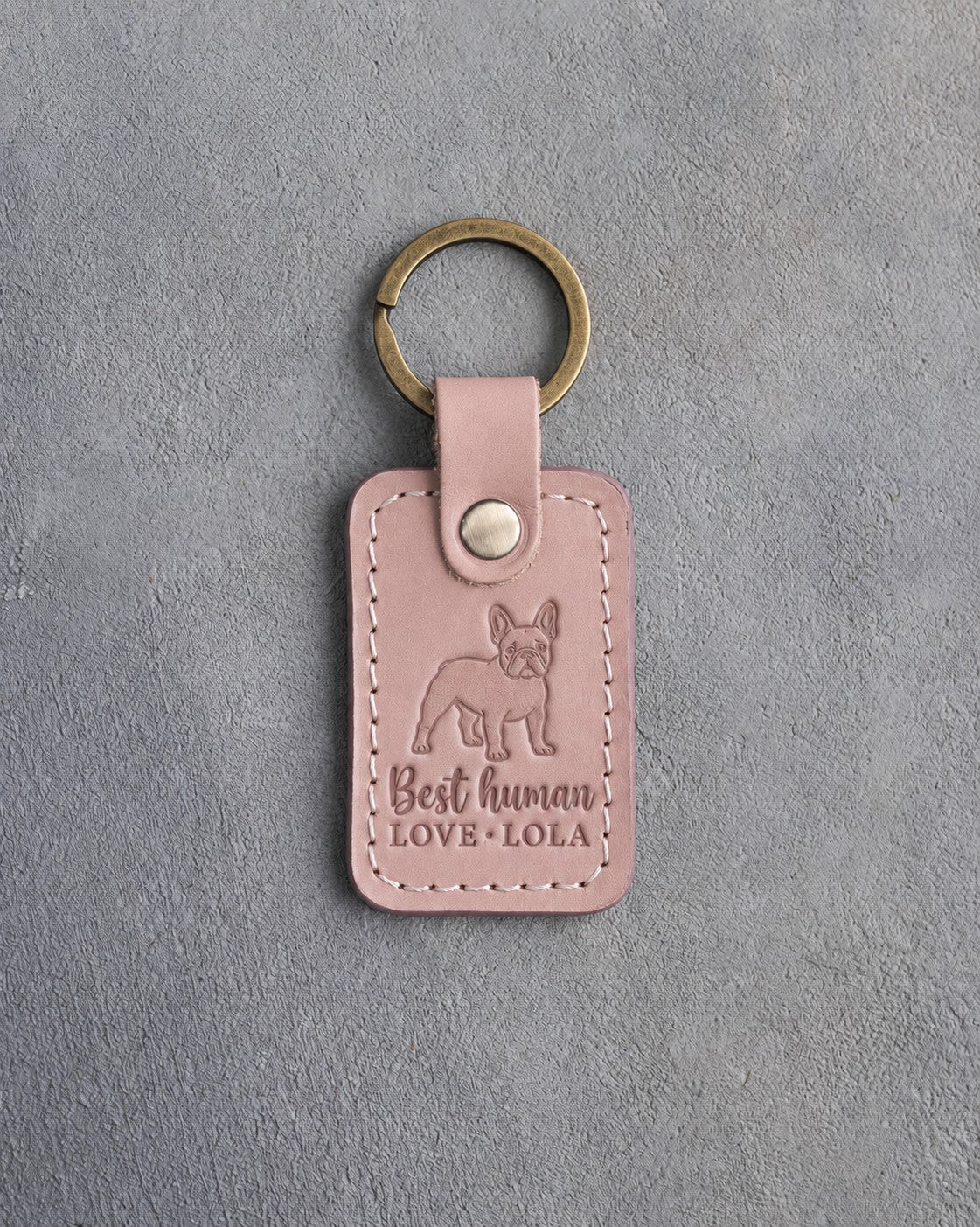 Blush pink leather keychain debossed with a French bulldog silhouette above Best human and LOVE · LOLA, on gray concrete