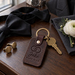 Jake BEST MAN keychain in dark brown leather on a walnut hotel dresser beside cufflinks and an untied black bow tie