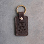 Dark espresso leather keychain debossed with two interlocked wedding rings above the name Jake and BEST MAN, on gray concrete