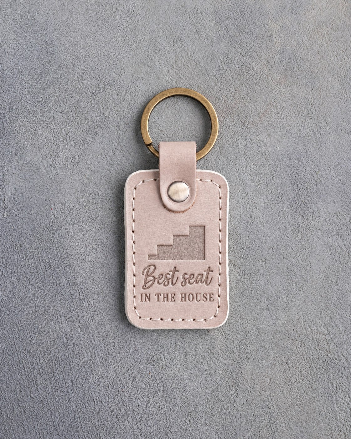 Gray sand leather keychain debossed with stepped bleachers and the words Best seat IN THE HOUSE, on gray plaster