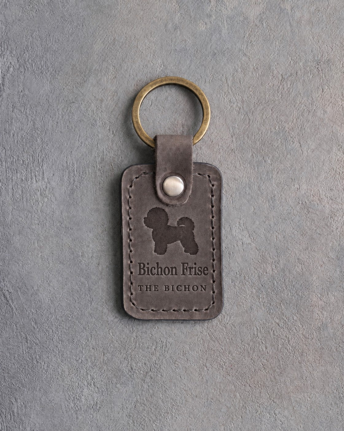 Country gray leather keychain debossed with a powder-puff Bichon Frise above Bichon Frise and THE BICHON, on gray concrete