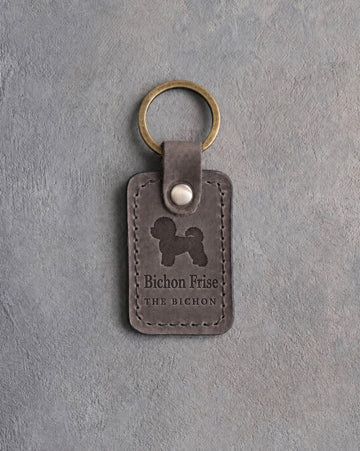 Country gray leather keychain debossed with a powder-puff Bichon Frise above Bichon Frise and THE BICHON, on gray concrete