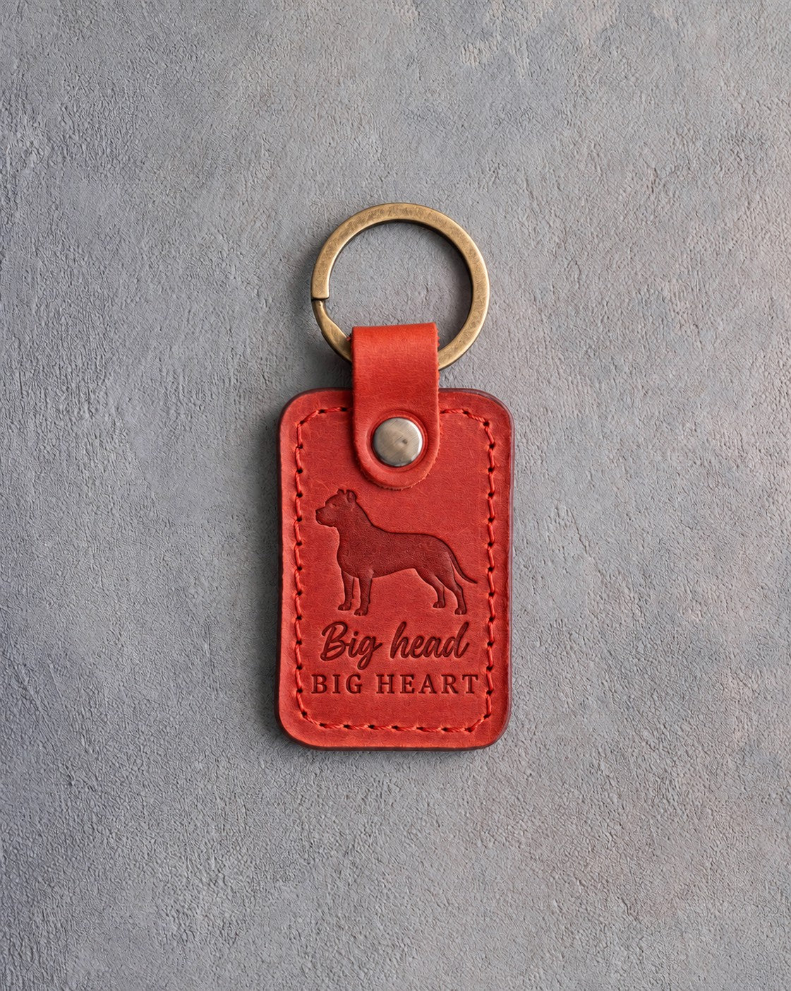 Poppy red leather keychain debossed with an American Staffordshire Terrier silhouette and the words Big head BIG HEART, on gray concrete