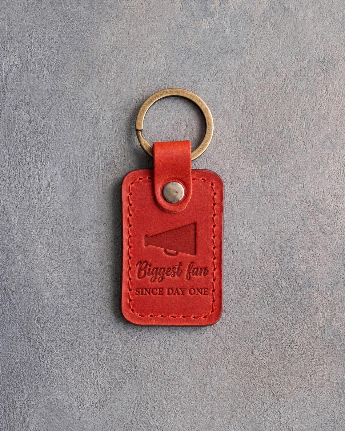 Poppy red leather keychain debossed with a megaphone and the words Biggest fan SINCE DAY ONE, on gray plaster