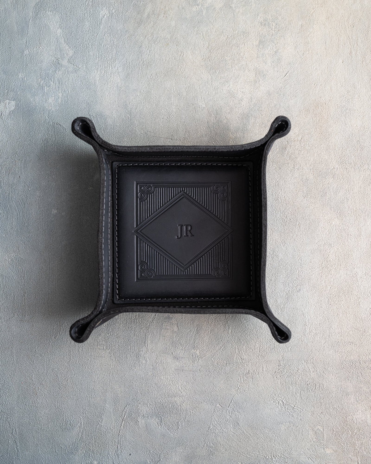 Black leather valet tray with snapped corners and the initials JR pressed into the diamond frame, seen from above