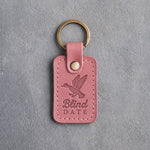 Rose pink leather keychain debossed with a mallard cupped to land above the words Blind DATE, on gray plaster