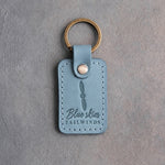 Turquoise leather keychain debossed with a two-blade propeller above the words Blue skies and TAILWINDS, on gray plaster