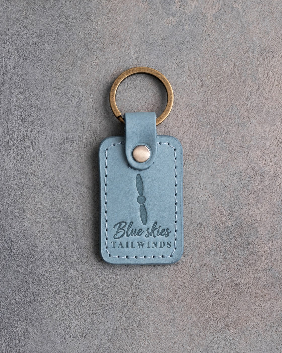 Turquoise leather keychain debossed with a two-blade propeller above the words Blue skies and TAILWINDS, on gray plaster