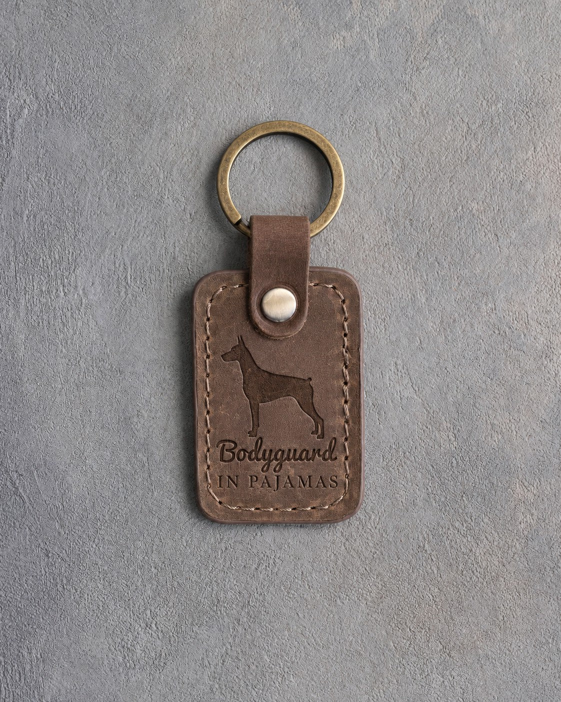 Sicilian brown leather keychain debossed with a Doberman silhouette and the words Bodyguard IN PAJAMAS, on gray concrete