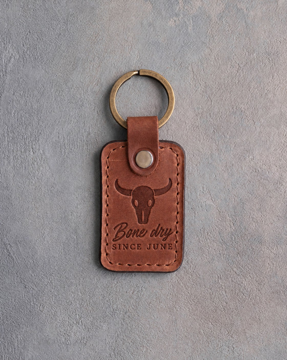 Arizona tan leather keychain debossed with a steer skull above Bone dry and SINCE JUNE, on gray plaster