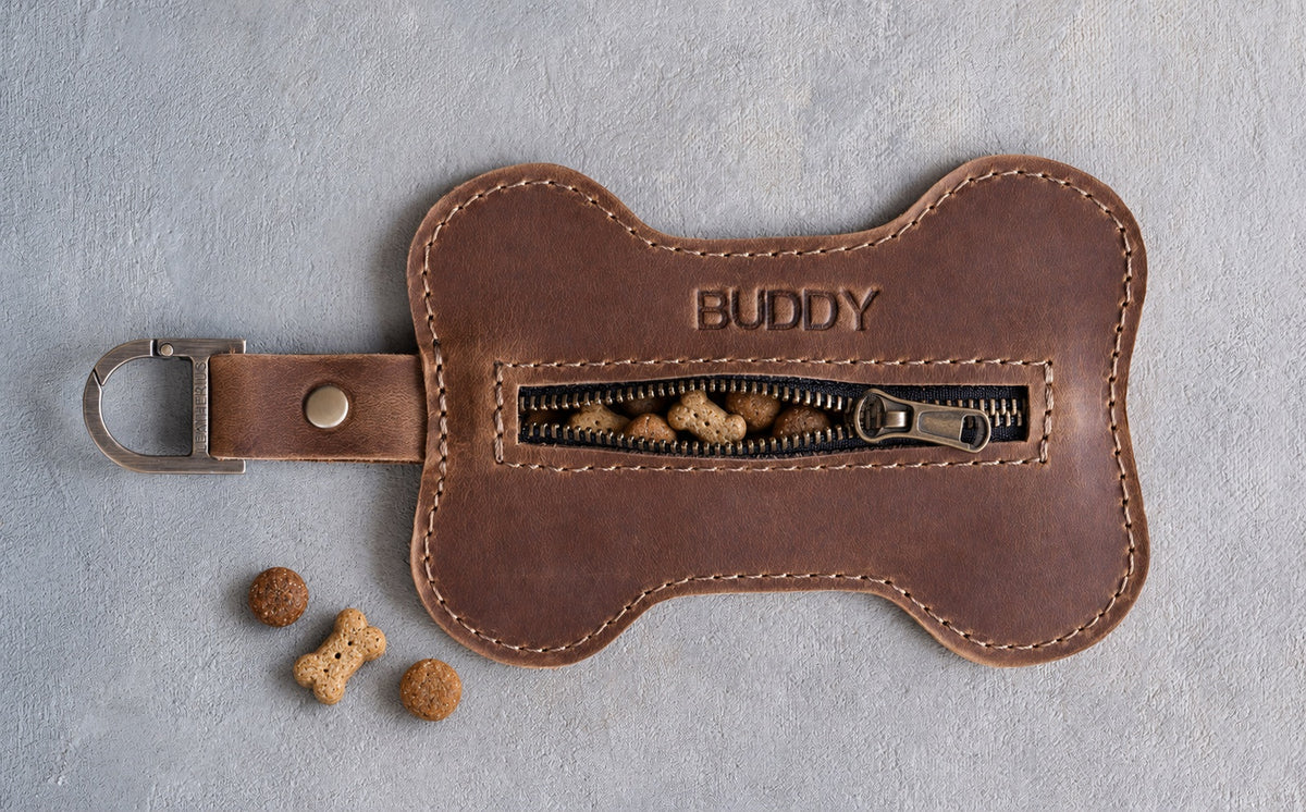 Brown bone shaped leather treat pouch stamped BUDDY, zipper slightly open with treats peeking out