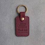 Sangria wine red leather keychain debossed with a stack of books above Book club and FOR THE WINE, on gray concrete