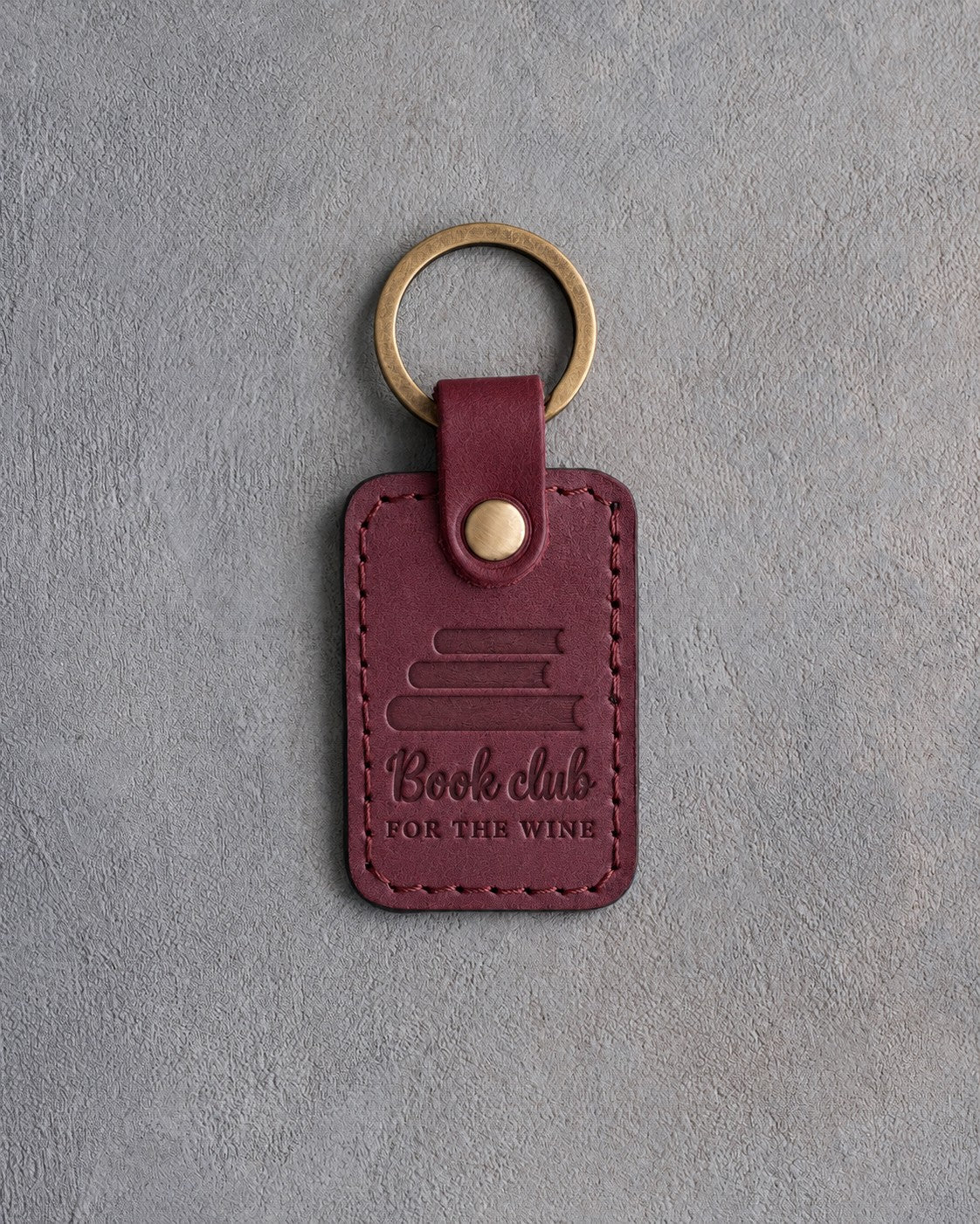 Sangria wine red leather keychain debossed with a stack of books above Book club and FOR THE WINE, on gray concrete