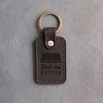 Dark espresso leather keychain debossed with an open book above Book was and BETTER, on gray concrete