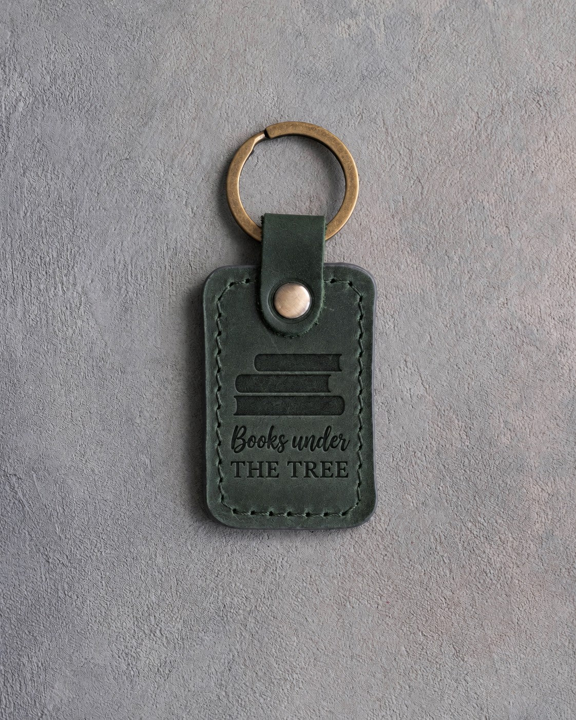 Forest green leather keychain debossed with a stack of books above Books under and THE TREE, on gray concrete