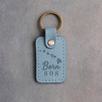 Turquoise leather keychain debossed with the Hawaiian island chain above Born and 808, on gray concrete