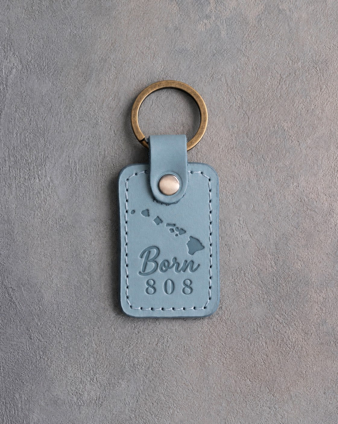 Turquoise leather keychain debossed with the Hawaiian island chain above Born and 808, on gray concrete