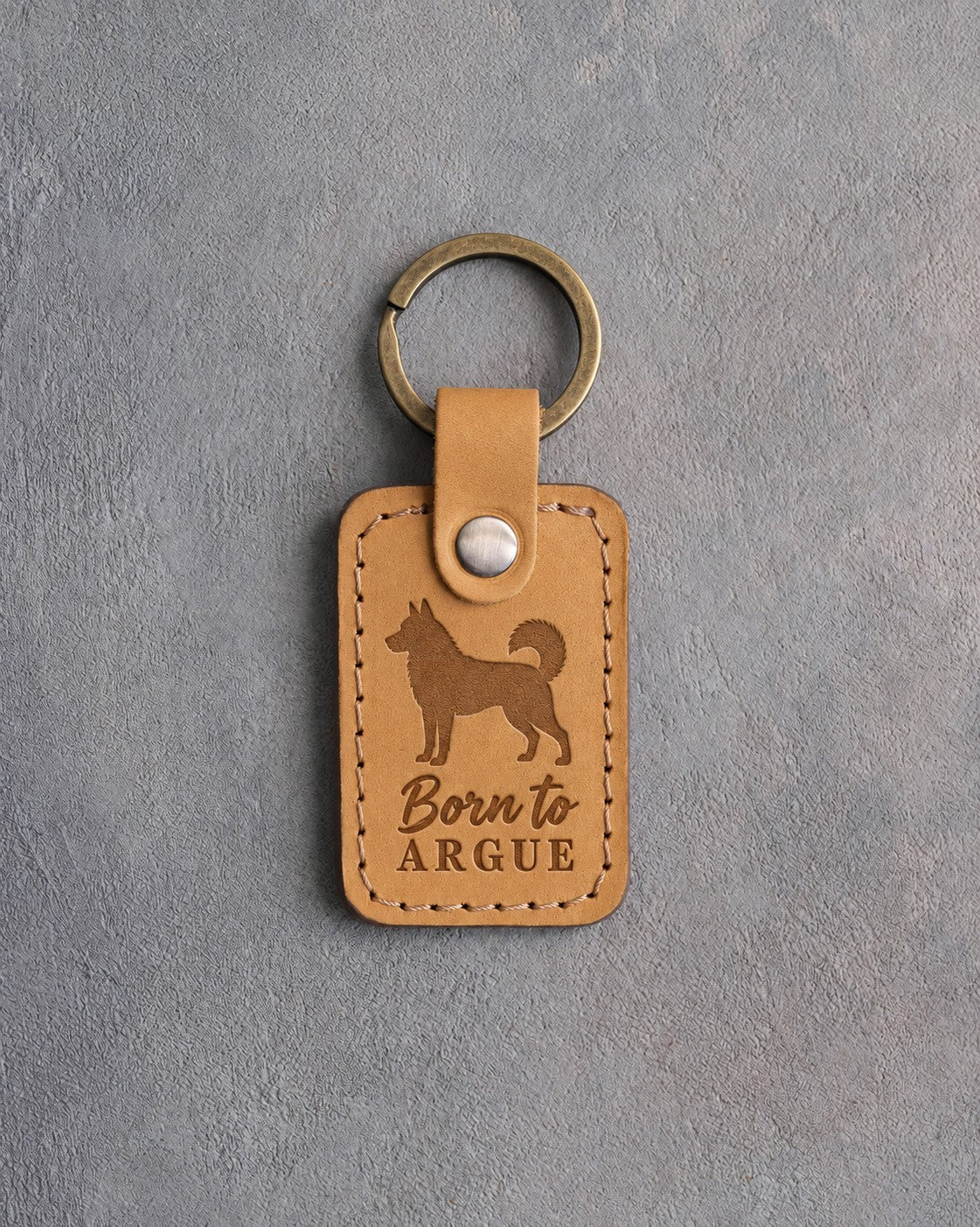 Indian Summer tan leather keychain debossed with a Siberian Husky silhouette and the words Born to ARGUE, on gray concrete
