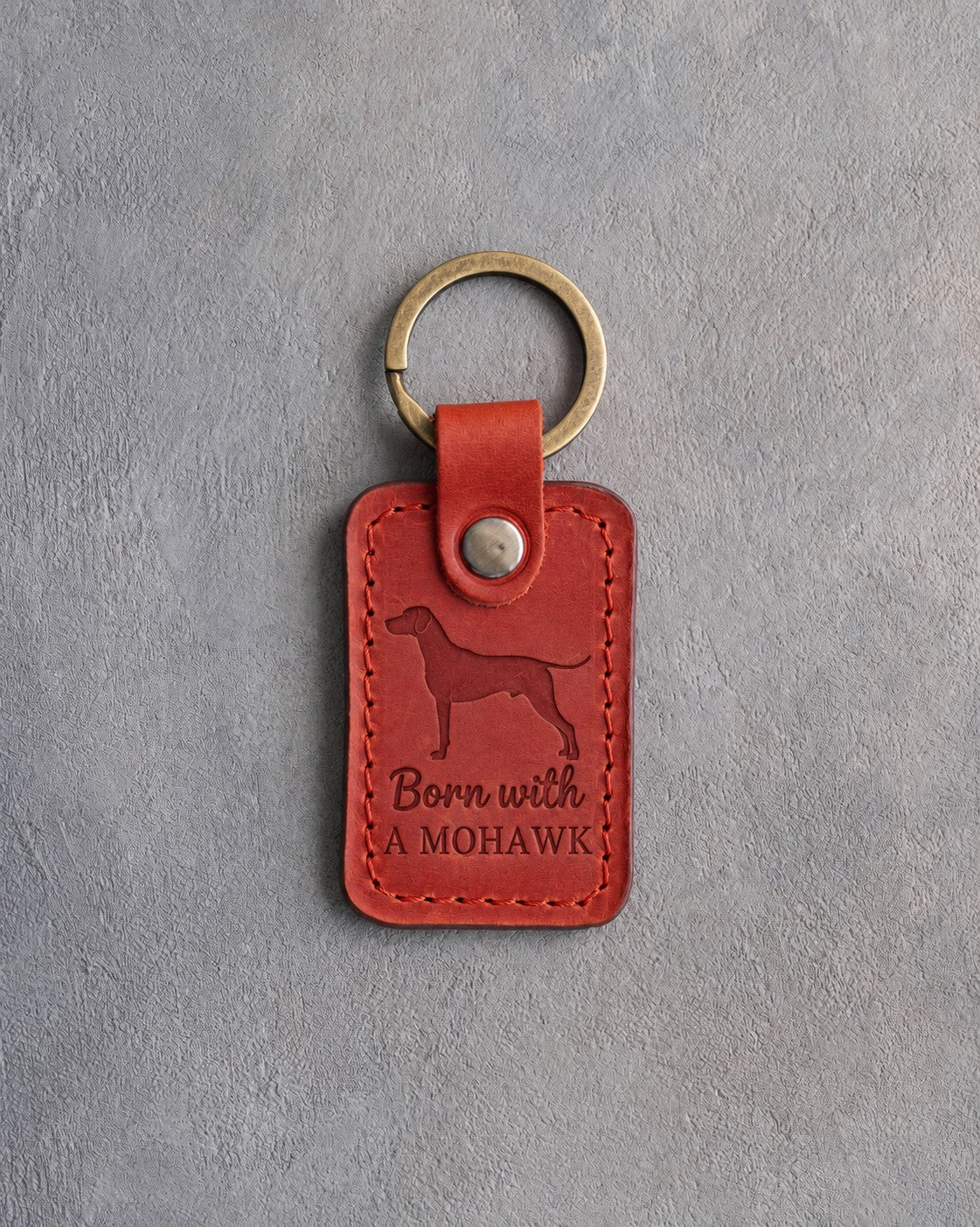 Poppy red leather keychain debossed with a Rhodesian Ridgeback silhouette and the words Born with A MOHAWK, on gray concrete