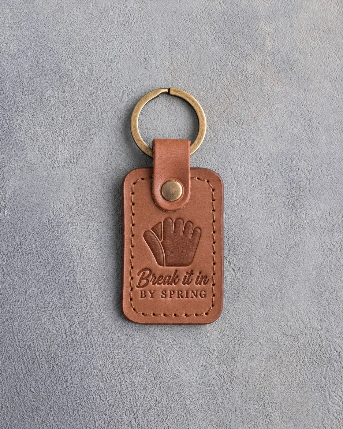 Caramel leather keychain debossed with a fielder's glove and the words Break it in BY SPRING, on gray plaster