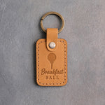 Golden tan leather keychain debossed with a golf ball on a tee above Breakfast BALL, on gray plaster
