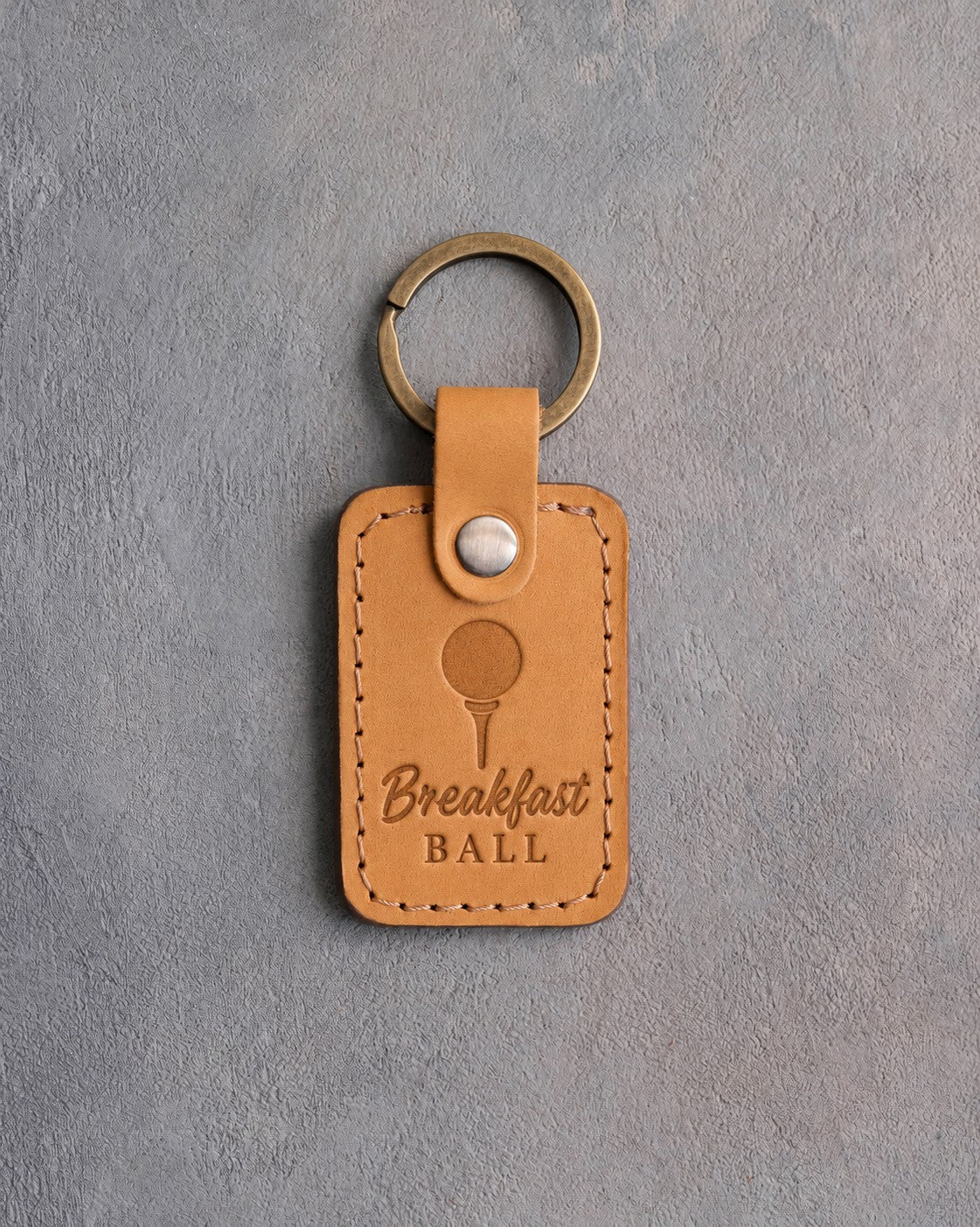 Golden tan leather keychain debossed with a golf ball on a tee above Breakfast BALL, on gray plaster