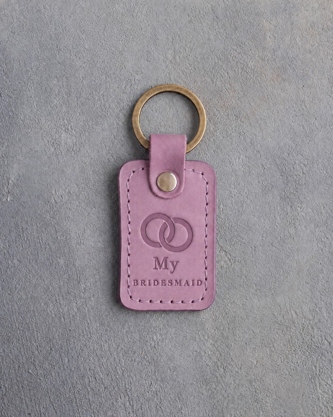 Lilac leather keychain debossed with two interlocked wedding rings above My and BRIDESMAID, on gray concrete