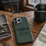 Forest green Travis BROOK TROUT iPhone case in full-grain leather on a knotty pine cabin table beside an open fly box and a green enamel coffee mug