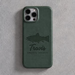 Back of a forest green leather iPhone case debossed with a brook trout above Travis and BROOK TROUT below the camera, on gray plaster