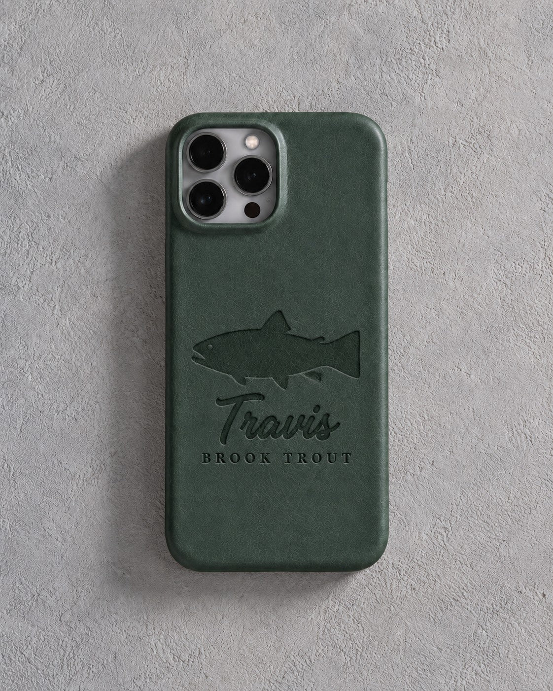 Back of a forest green leather iPhone case debossed with a brook trout above Travis and BROOK TROUT below the camera, on gray plaster