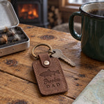 Sicilian brown Brookie DAD keychain in full-grain leather on a knotty pine cabin table beside an open fly box and a green enamel coffee mug