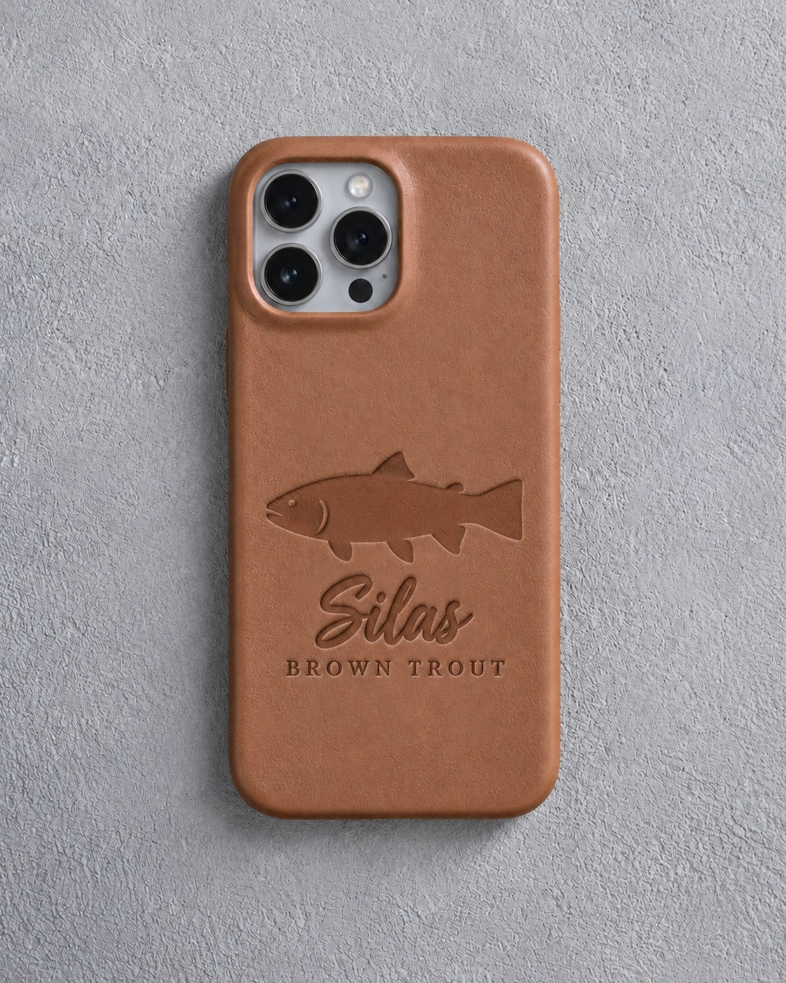 Back of a caramel leather iPhone case debossed with a brown trout above Silas and BROWN TROUT below the camera, on gray plaster