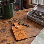 Indian Summer Silas BROWN TROUT keychain in full-grain leather on a knotty pine cabin table beside an open fly box and a green enamel coffee mug