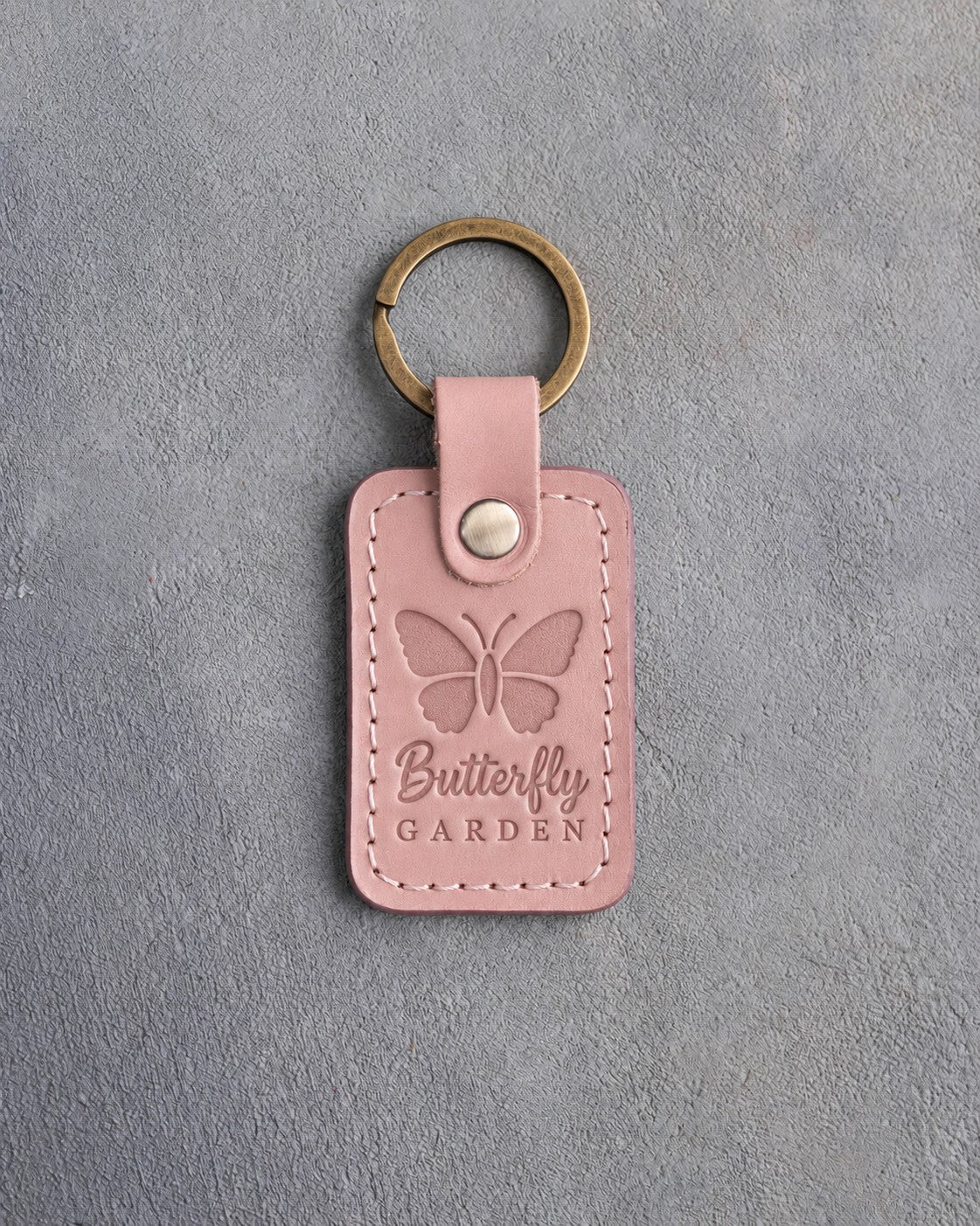 Blush pink leather keychain debossed with a butterfly above Butterfly GARDEN, on gray concrete