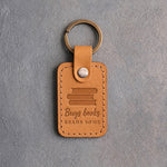 Indian Summer tan leather keychain debossed with a pile of books above Buys books and READS SOME, on gray concrete